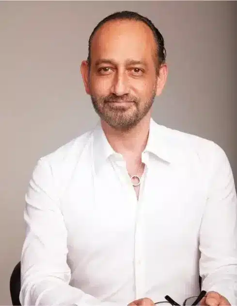 A middle-aged cosmetic surgeon with a beard, smiling slightly at the camera, wearing a white shirt and holding glasses.